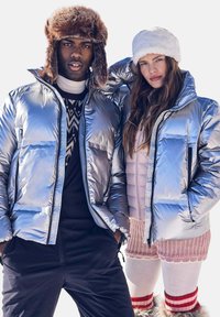 Giubbotti imbottiti metallici argento con grandi tasche frontali. Un modello indossa un berretto in pelliccia finta; l'altro indossa un cappello bianco in pelliccia e maglioni sovrapposti.