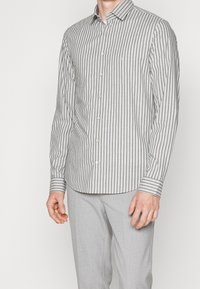 Witte overhemd met dunne zwarte verticale strepen, lange mouwen en een standaard kraag. Heeft een afgeronde zoom en contrasterende manchetten met strepen.