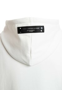 Sudadera blanca con tejido texturizado, una capucha grande y una etiqueta negra con texto blanco y botones a presión en la zona del cuello.