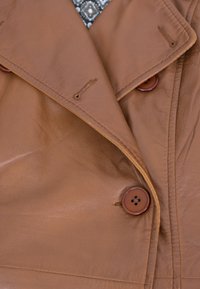 Brauner Ledertrenchcoat mit einer doppelt geknöpften Vorderseite, ausgestattet mit zwei runden braunen Knöpfen und einer glatten Textur. Gemustertes Innenfutter sichtbar.