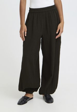 Person trägt lockere schwarze, geknitterte Hosen mit elastischen Bündchen und Taillenbund, kombiniert mit schwarzen Slip-On-Schuhen und einem weißen Oberteil.