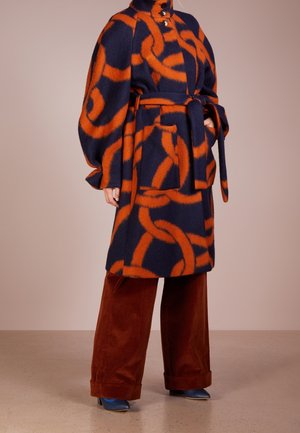 Personne portant un manteau ceinturé bleu marine et orange à motifs avec de grandes poches, associé à un pantalon large couleur rouille et des chaussures pointues bleues.