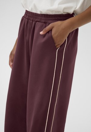 Personne portant un pantalon ample bordeaux foncé avec un passepoil blanc sur le côté, une ceinture élastique et une main reposant dans la poche.