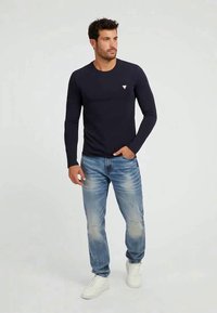Homem vestido com uma camisa preta ajustada de manga longa, jeans claros desgastados e ténis brancos, de pé com uma mão no bolso.