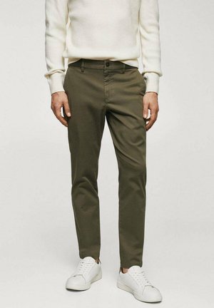 Homme portant un pantalon slim vert olive, un pull blanc et des baskets blanches, debout devant un fond clair uni.
