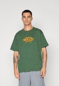Grøn bomulds T-shirt med en slidt tekstur, med et gult Nike-logo omkranset af et tornet design. Korte ærmer, afslappet pasform.