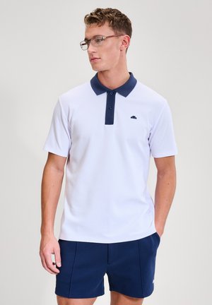 Mann trägt ein weißes Poloshirt mit marineblauem Kragen und Knöpfen, marineblaue Shorts und eine Brille, steht mit einer Hand in der Tasche.