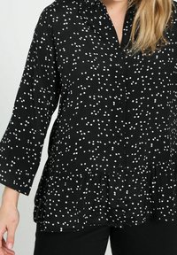 Blusa negra con lunares blancos, con botones en el frente, cuello y dobladillo acampanado. Mangas largas de corte holgado y tela ligeramente texturizada.