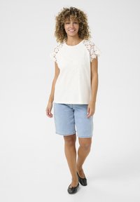 Weißes kurzärmeliges Oberteil mit Spitzenakzenten an den Schultern, kombiniert mit hellblauen Denim-Shorts. Das Oberteil hat einen runden Ausschnitt und eine lockere Passform.