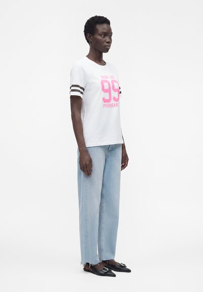 Γυναίκα που φοράει ένα λευκό T-shirt με ροζ «99» και ρίγες στα μανίκια, ανοιχτό μπλε τζιν παντελόνι με φαρδιά μπατζάκια και μαύρες μυτερές μπαλαρίνες με αγκράφες.