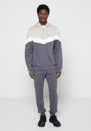 Uomo in piedi che indossa una felpa con cappuccio con zip color-block e pantaloni della tuta grigi coordinati con scarpe da ginnastica nere su uno sfondo chiaro e semplice.