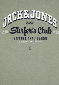 Camiseta de algodón verde con texto grande en blanco y navy que dice "JACK & JONES Surfer's Club", decorada con olas y tablas de surf cruzadas abajo.