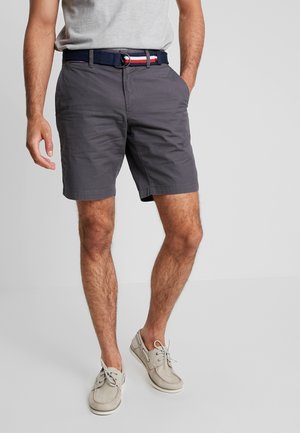Graue Baumwollshorts mit Seitentaschen, ein navyblauer Gürtel mit roten und weißen Akzenten, kombiniert mit hellgrauen Boots.