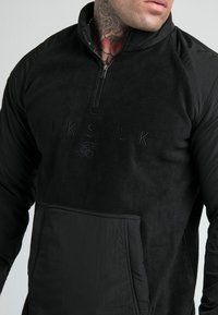 Felpa nera con zip e collo alto, in tessuto polar, logo ricamato sul davanti e tasca a kangaroo; maniche in contrasto con finitura liscia.