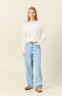 Maglione bianco a maglia con motivo a onde colorate, abbinato a jeans azzurri a vita alta. La modella indossa pantofole beige e orecchini semplici.