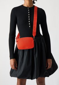 Červená křížová taška z texturované kůže obdélníkového tvaru s nastavitelným popruhem s logem, doplněná o černé žebrované šaty a sukni.
