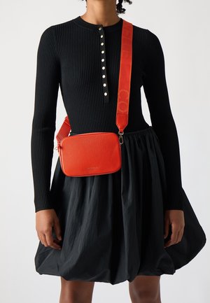 Red textured leather crossbody bag with a rectangular shape, adjustable strap featuring a logo, paired with a black ribbed dress and skirt.