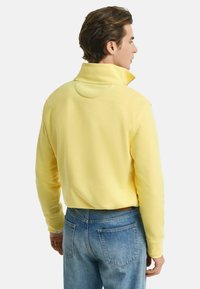 Gele fleece trui met een hoge kraag, ribgebreide boorden en een relaxte pasvorm. Gepaard met lichtblauwe spijkerbroeken, waarbij de details aan de achterkant zichtbaar zijn.
