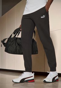 Zwarte joggers met zijzakken en een wit Puma-logo, gecombineerd met zwart-witte sneakers met rode accenten en een zwarte sporttas.