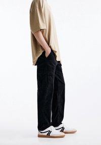 Béžové bavlněné oversized tričko, černé cargo kalhoty s kapsami a bílé tenisky s černými akcenty a gumovou podrážkou. Minimalistický design.