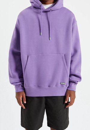Persoon die een paarse hoodie met een voorzak en zwarte korte broek draagt, staand tegen een effen witte achtergrond.