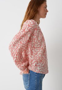 Blusa leggera e stampata di bianco con onde rosse. Presenta maniche lunghe e svasate e una vestibilità rilassata, abbinata a jeans blu.