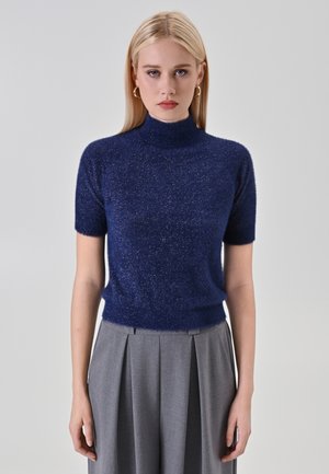 Blonde Frau, die einen glitzernden marineblauen kurzärmeligen Rollkragenpullover und hoch taillierte graue Plisseehosen trägt, steht vor einem schlichten Hintergrund.