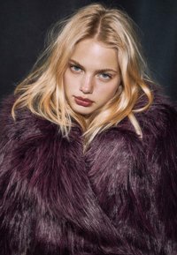 Blondynka z niebieskimi oczami, ubrana w dużą, ciemnofioletową sztuczną futrzaną kurtkę na ciemnym tle, patrzy bezpośrednio w kamerę.