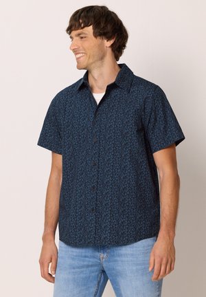 Mann lächelt und schaut nach links, trägt ein dunkelblaues kurzärmeliges Hemd mit einem dezenten Blumenmuster und hellblaue Jeans.
