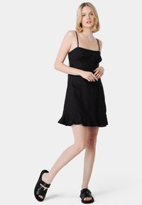 Abito di lino nero con spalline sottili, corpino aderente e orlo arricciato. Abbinato a sandali neri a infradito. Design semplice ed elegante.