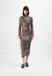 Woman standing wearing a long-sleeve leopard print midi dress, black pointed shoes, a pearl necklace, and holding a small pale pink handbag.