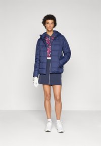 Veste matelassée marine avec capuche, dotée d'un design matelassé et de poignets élastiques, portée sur un haut à motifs et associée à une jupe marine.