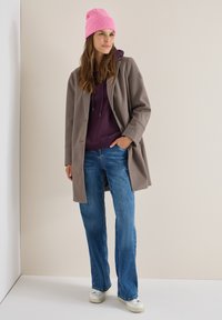 Abrigo de lana gris sobre una sudadera morada, jeans azules y zapatillas blancas. Gorro de punto rosa en la cabeza, corte relajado con piernas rectas.