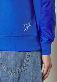 Blå sweatshirt med texturerade ärmar, med en vit broderad logotyp och signatur vid nederkanten. Bärs med ljusblå denimjeans.