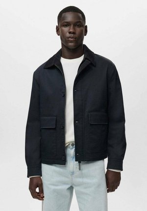 Black cotton jacket with a corduroy collar, two front pockets, and a button closure. Worn over a white ribbed sweater and light jeans.