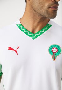 Bijela nogometna majica s zelenim akcentnim uzorkom, koja sadrži crveni Puma logo i okrugli marokanski grb s zlatnim i zelenim detaljima.