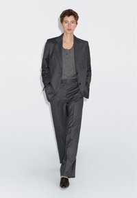 Grau maßgeschneiderter Anzug mit einem Ein-Knopf-Blazer, passenden Stoffhosen und einer grauen Bluse, die eine glatte Textur und eine klassische Passform aufweist. Schwarze Slipper.