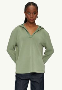 Groene losse hoodie met een V-hals en koord, gemaakt van zachte stof; gecombineerd met donkere spijkerbroek en met lange mouwen.