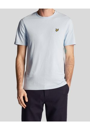 Mand iført en lyseblå kortærmet t-shirt med en lille gul fuglelogo på brystet og mørke bukser mod en grå baggrund.