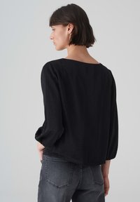 Schwarze, locker sitzende Bluse mit weitem Ausschnitt, langen Puffärmeln und einem leicht abgerundeten Saum. Der Stoff wirkt weich und hat einen dezenten Glanz.