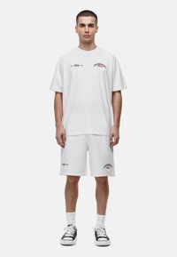 Witte katoenen t-shirt en korte broek set; voorzien van bedrukte tekst en accenten in het rood. Compleet met zwarte sneakers en witte sokken.