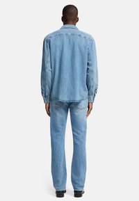 Camicia di denim azzurro chiaro con maniche lunghe e polsini abbottonati, abbinata a jeans dritti azzurro chiaro, vista da dietro.