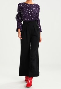 Blusa viola con pois bianchi, maniche a campana, abbinata a pantaloni a gamba larga neri a vita alta e stivaletti rossi lucidi.