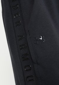 Black athletic pants featuring a textured side stripe with "UNDER ARMOUR" in glossy lettering and a small reflective logo on the leg.