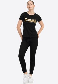 Melnā t-kreklā ar īsām piedurknēm, uz kura redzams zelta Lonsdale London logo, apvienots ar melnām skinny džinsa biksēm un baltām sporta apavām. Vienkārša dizaina un materiāli.