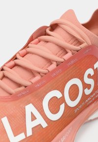 Αθλητικό παπούτσι σε ανοιχτό ροζ με υφή στο επάνω μέρος, έντονο λευκό λογότυπο "LACOSTE" και λείες κορδόνια. Το υλικό φαίνεται συνθετικό.