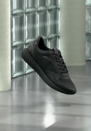 Baskets noires de la marque BOSS avec des détails en mesh et cuir, flottant au-dessus d'un sol poli contre des murs en blocs de verre.