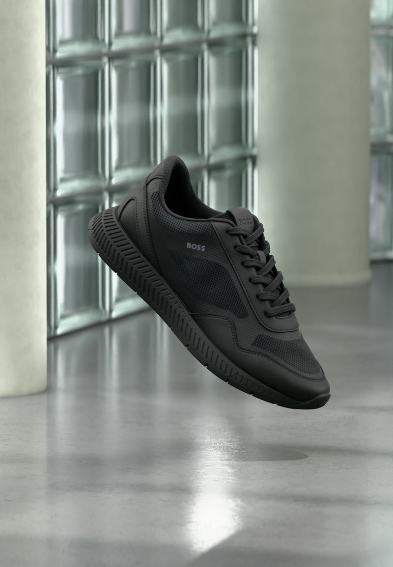 Baskets noires de la marque BOSS avec des détails en mesh et cuir, flottant au-dessus d'un sol poli contre des murs en blocs de verre.