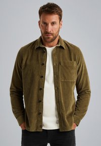 Olijfgroene corduroy overhemdjasje met knoopsluiting, een borstzak en gestructureerde stof. Ggedragen over een witte trui.