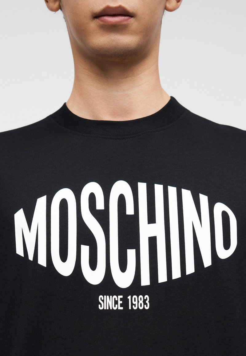 T-shirt noir à col rond avec le texte blanc audacieux « MOSCHINO » et « SINCE 1983 » dans une police moderne. Le tissu semble lisse avec une coupe classique.